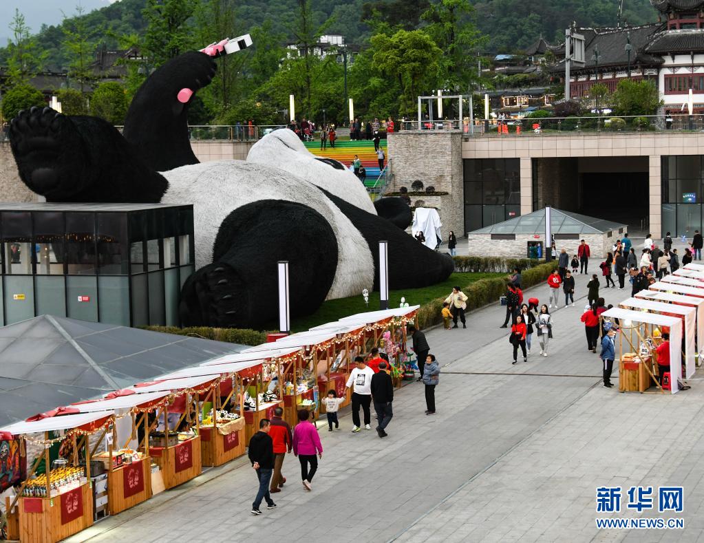 　　4月7日，在四川省都江堰市仰天窝广场，游客在“自拍熊猫”雕塑旁边的文创集市消费。　　近日，由荷兰艺术家弗洛伦泰因·霍夫曼设计的大熊猫自拍艺术品雕塑在四川省都江堰市仰天窝广场与公众见面。这只仰卧翘脚自拍的“大熊猫”长26.5米、宽11米、高12米，重达130吨，由300万根漆面不锈钢丝固定而成。　　据了解，这只熊猫雕塑以2005年大熊猫闯入都江堰闹市区的故事为创作灵感。熊猫手持一部巨型智能手机，吸引市民和游客前来观赏，合影“打卡”。　　新华社记者 王曦 摄